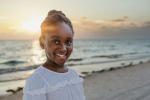 Young person in a white dress smiles brightly on a beach at sunset.