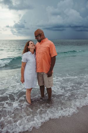 A couple stands in ocean waves as storm clouds gather on the horizon.