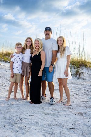 A family of five poses together on a sandy beach with sea grass and cloudy blue skies in the background.