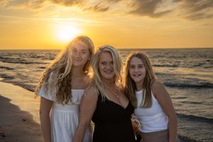 Three women pose together on a beach at sunset with waves crashing behind them.