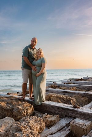 Maternity photo of couple standing on driftwood at beach during sunset.