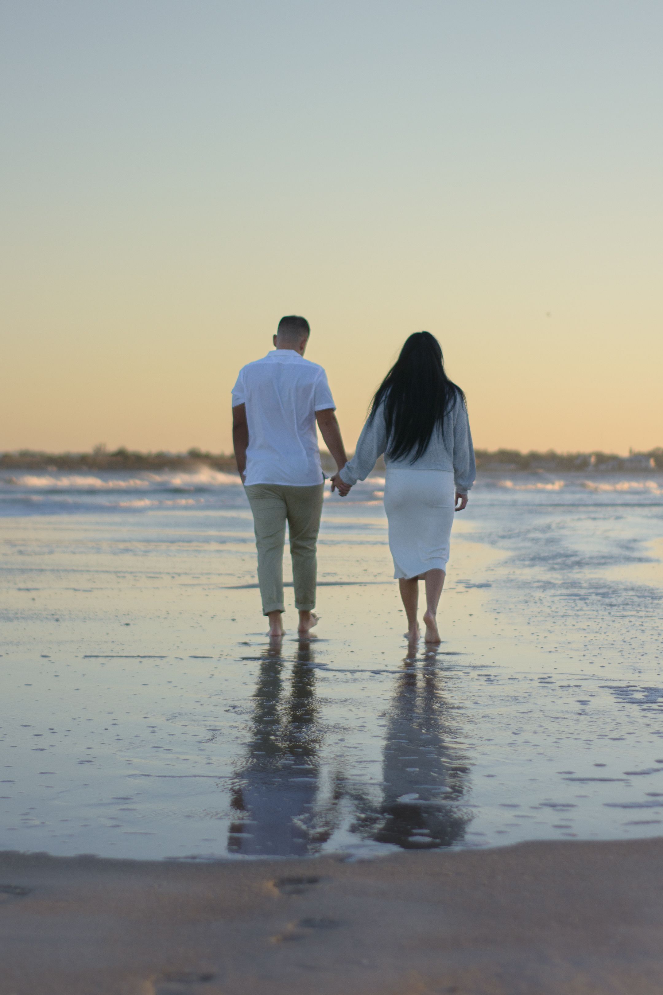A couple walks barefoot along the shoreline at sunset holding hands.