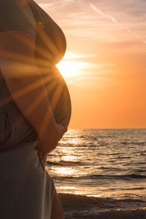 Silhouette of pregnant belly against dramatic orange sunset over ocean waves.