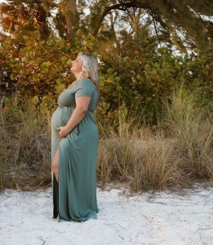 Expectant mother in teal dress poses in natural setting with autumn foliage.
