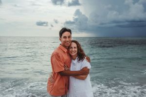 A couple embraces on the beach with stormy clouds and ocean waves in the background.