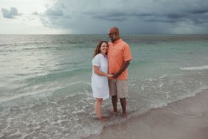 A couple stands together on a beach with stormy clouds overhead and turquoise waters in the background.