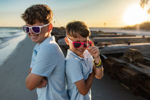 Two children wearing sunglasses pose playfully on a beach at sunset.