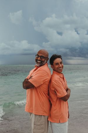 Two people in coral-colored shirts pose back-to-back on a beach with dramatic storm clouds.