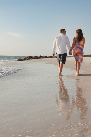 Couple walking hand in hand along a beach shoreline at sunset with waves and rocks in the background.