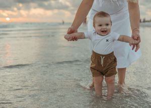 A baby in brown shorts takes first steps on wet beach sand during sunset.