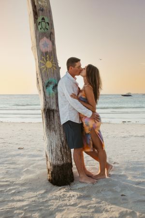 Romantic beach scene with couple embracing against a wooden pole during sunset with ocean views.