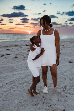 Two people in white dresses embrace while walking along a beach at sunset.