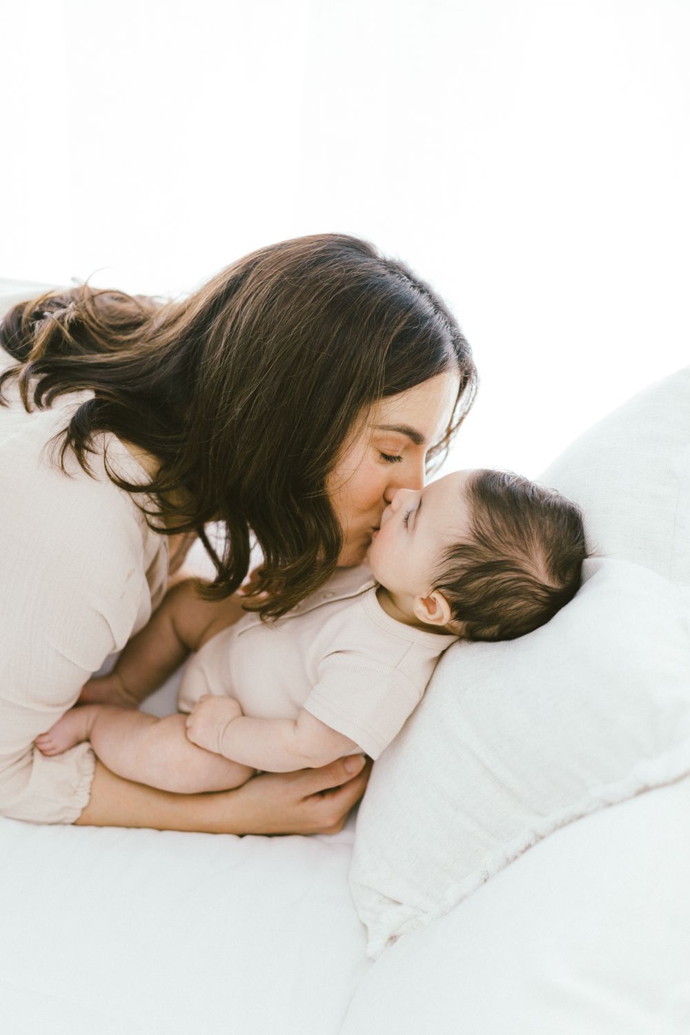 Loving parent leans in close to baby while resting together on white sheets.