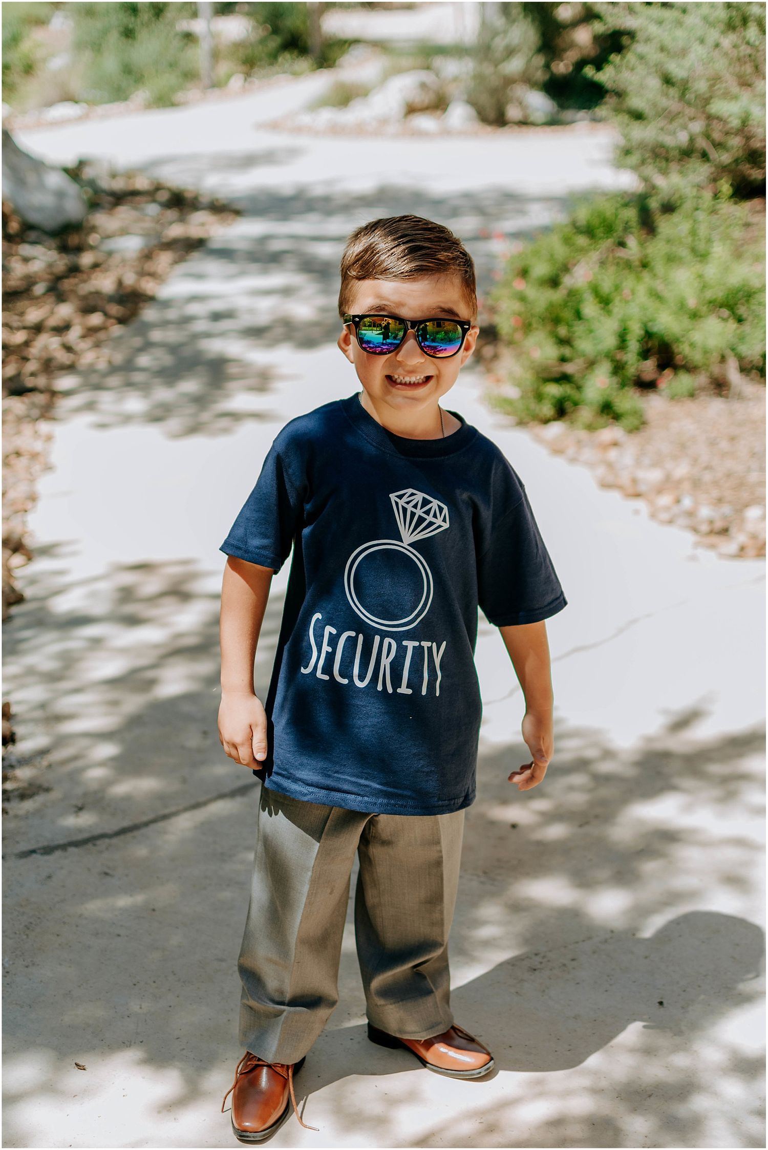 Young ring bearer poses in navy security t-shirt and sunglasses on outdoor path.
