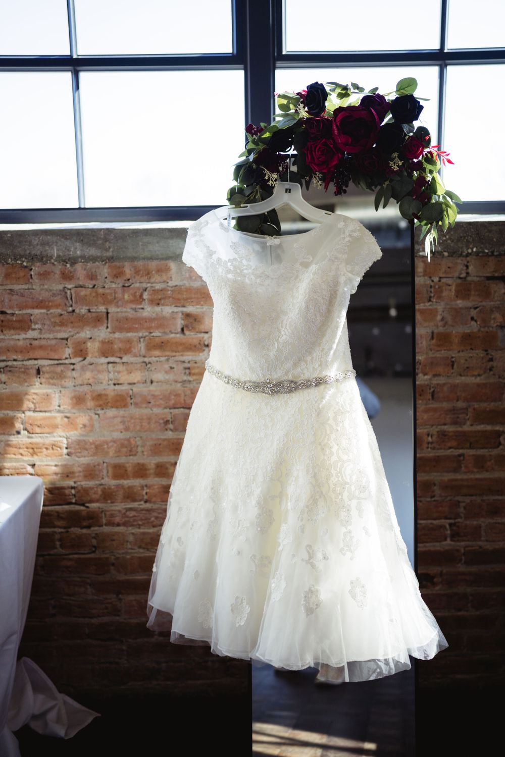A white lace wedding dress with cap sleeves and beaded belt hanging near exposed brick windows.