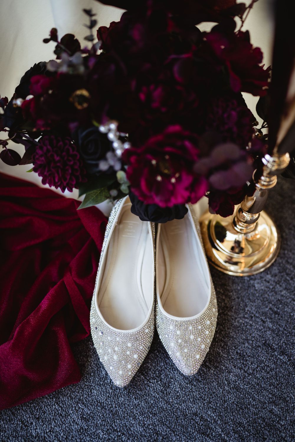 Sparkly flat wedding shoes next to deep burgundy flowers on grey carpet.