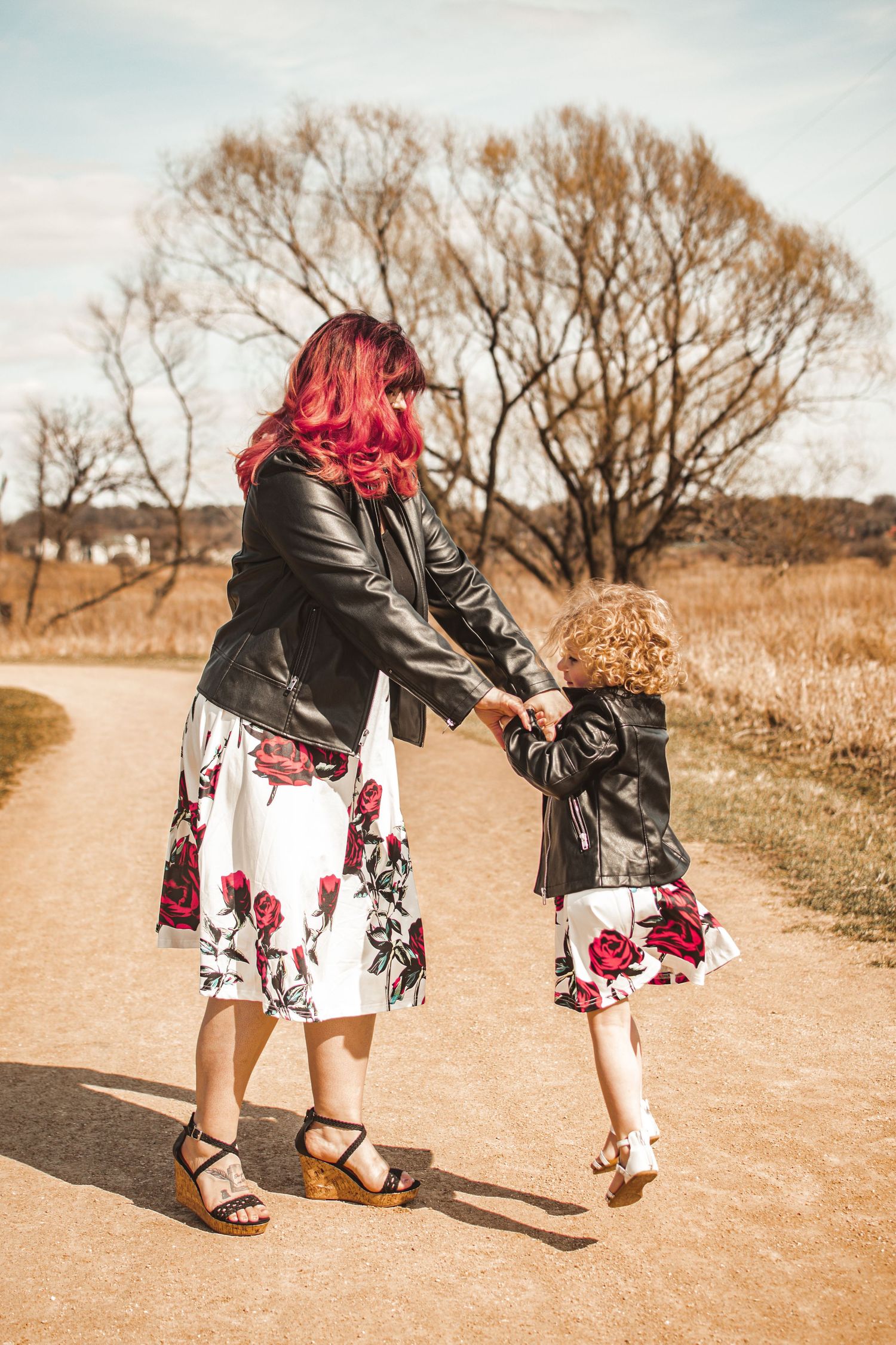 Two people in matching floral dresses and leather jackets hold hands while walking on a path.