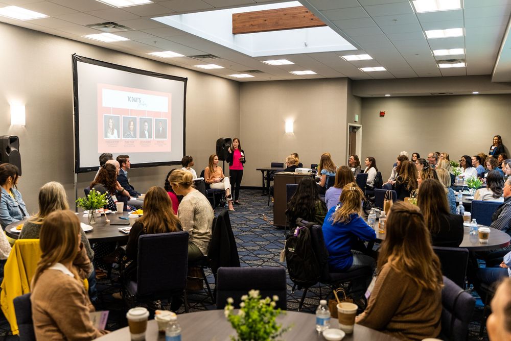 Conference room filled with attendees seated at round tables during presentation.