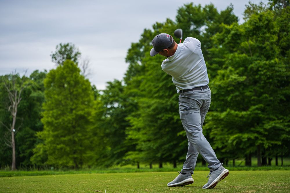 Golfer in grey attire mid-swing on golf course with forest backdrop.