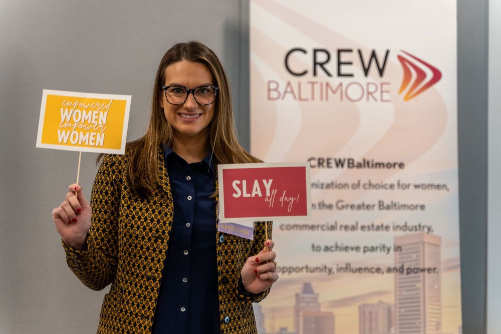Professional holding inspirational signs at CREW Baltimore networking event with organization banner in background.