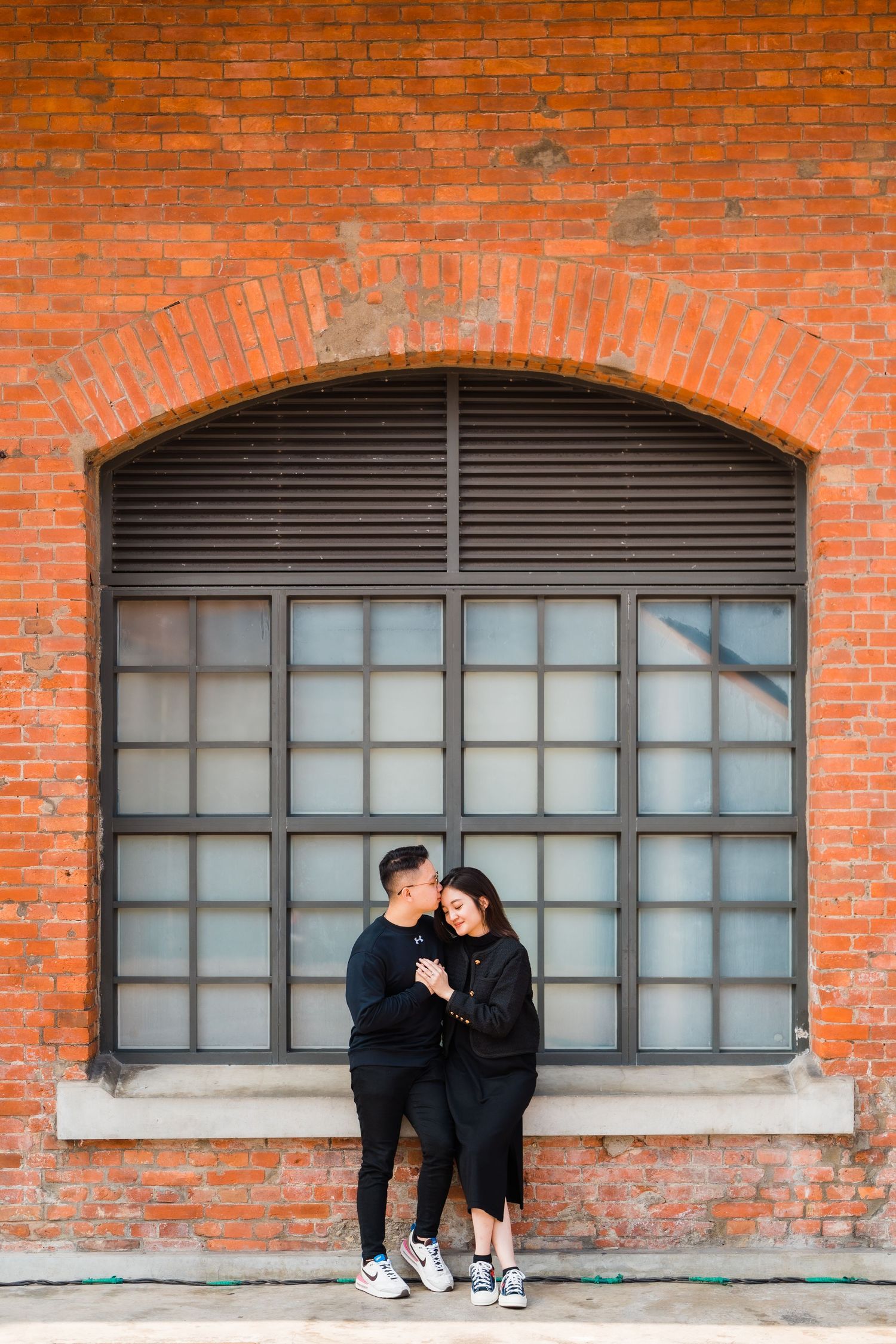 A couple in matching black outfits sharing an intimate moment against a rustic brick wall with arched window.