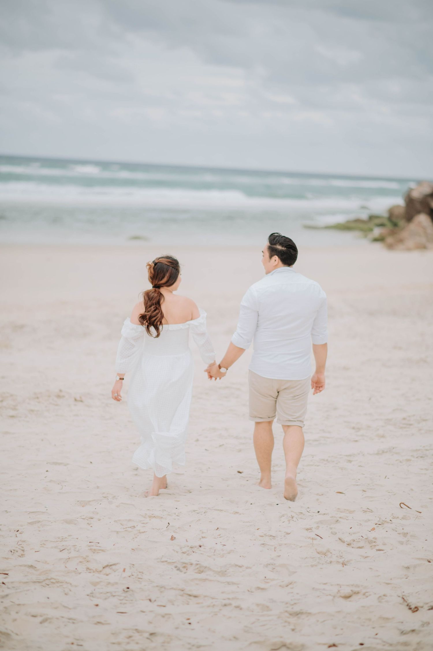 A couple walking hand in hand along a pristine beach with waves crashing in the background.