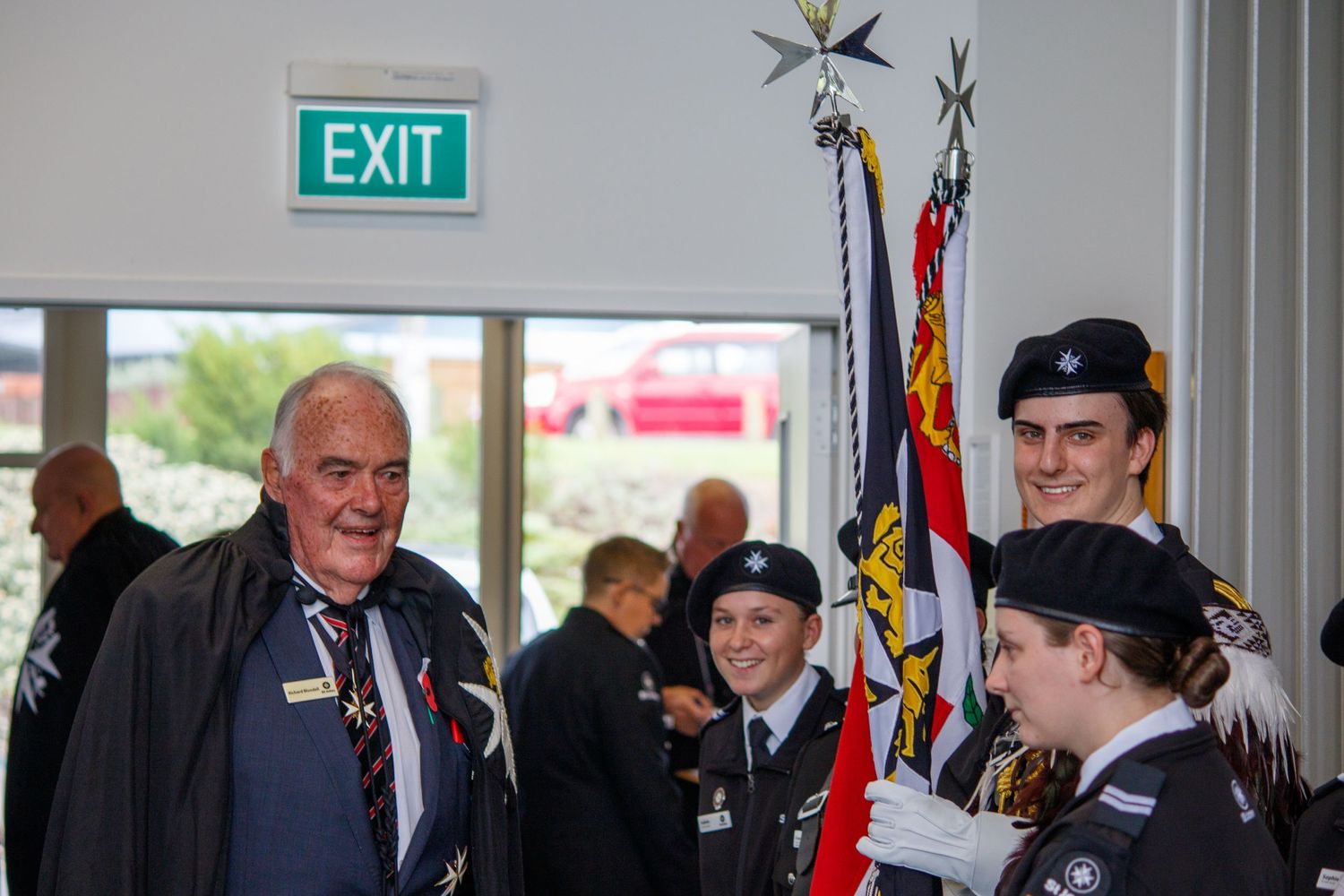 Military cadets holding ceremonial flags with veteran at formal indoor ceremony near exit sign.