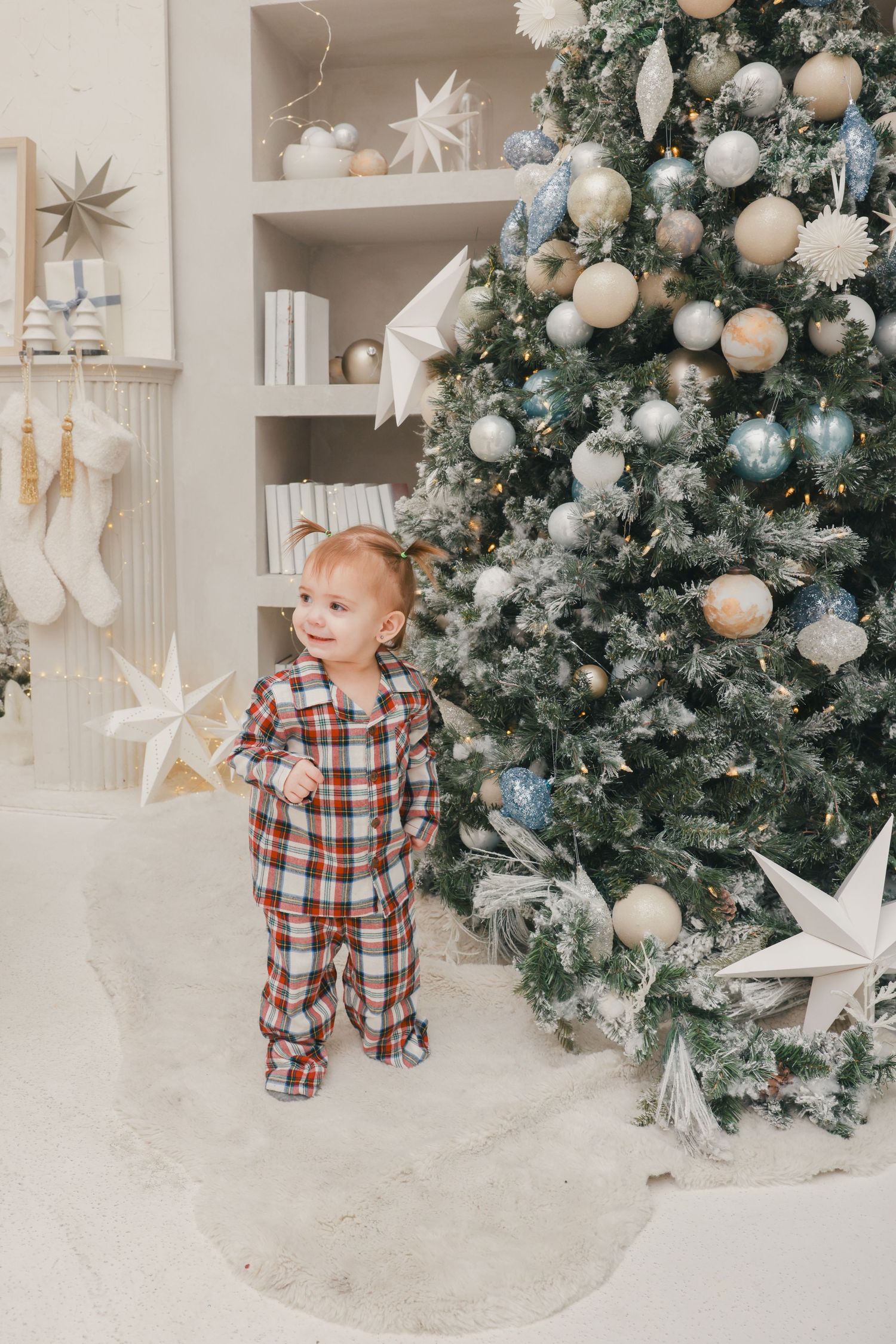 A toddler in plaid pajamas stands beside a decorated white Christmas tree with ornaments and stars.