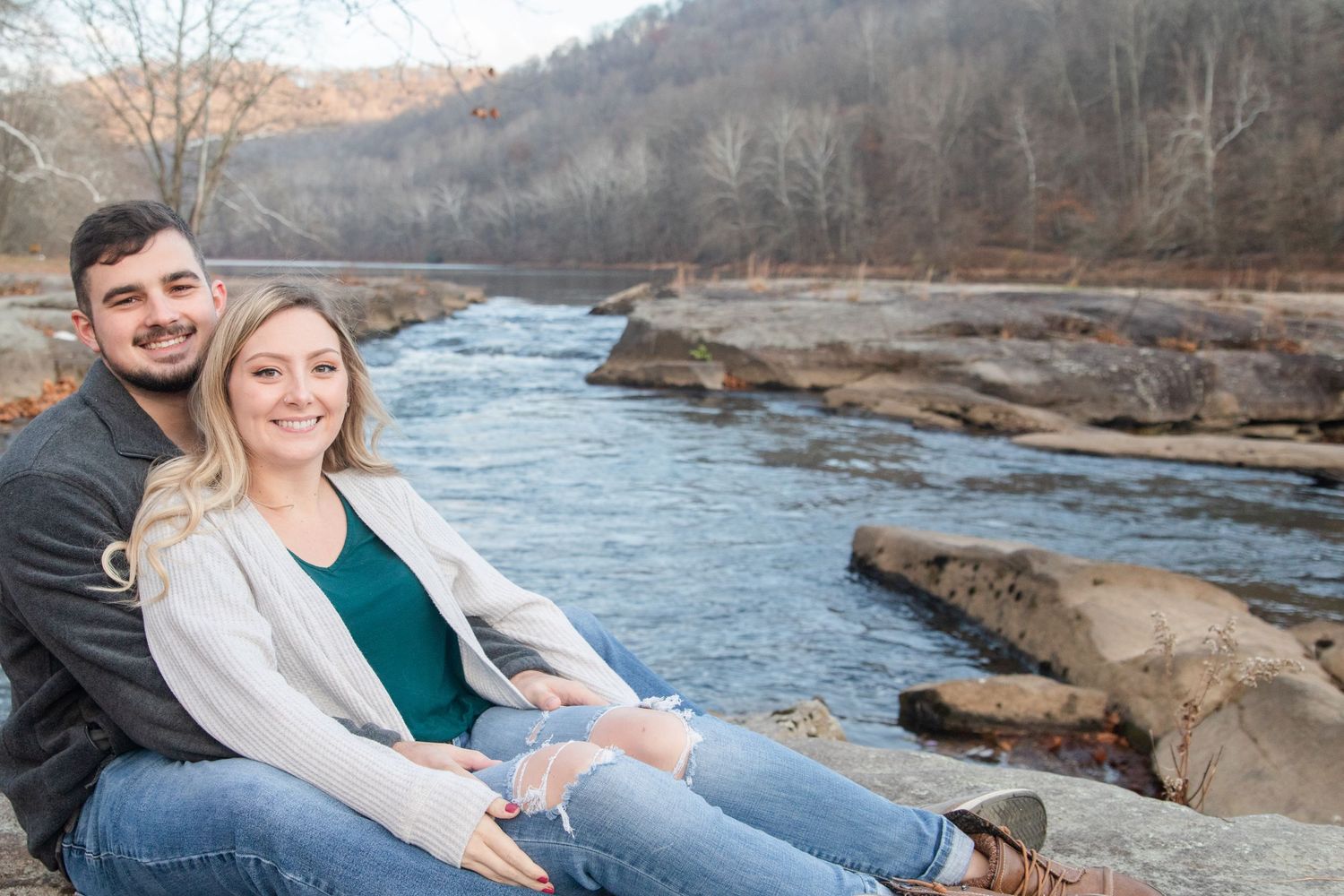 A couple sits together on rocks by a flowing river surrounded by autumn trees in a scenic outdoor setting.