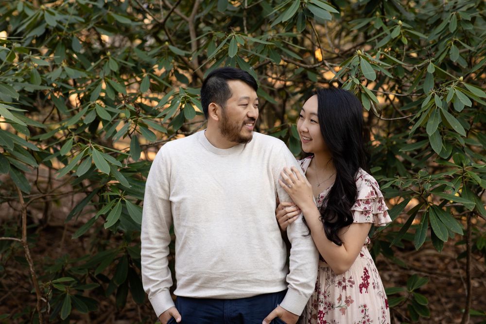 A couple laughs together while posing against green foliage in an outdoor setting.