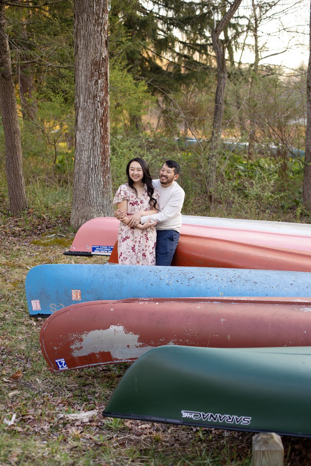 A couple sits together on colorful canoes stacked by the forest's edge.