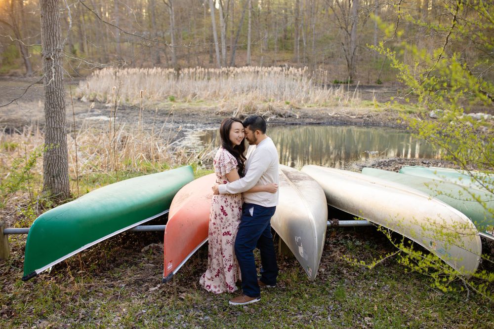 Couple embraces by a lake with stacked canoes in autumn scenery.