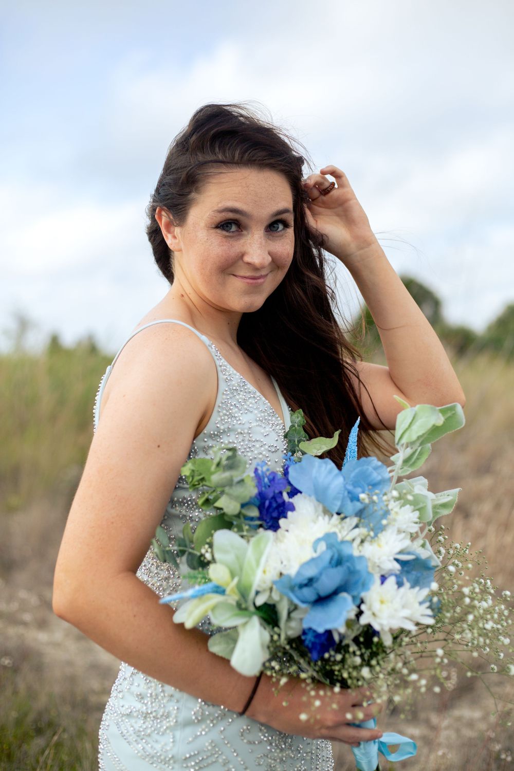 Bride in sequined dress holding blue and white bouquet in outdoor field setting.