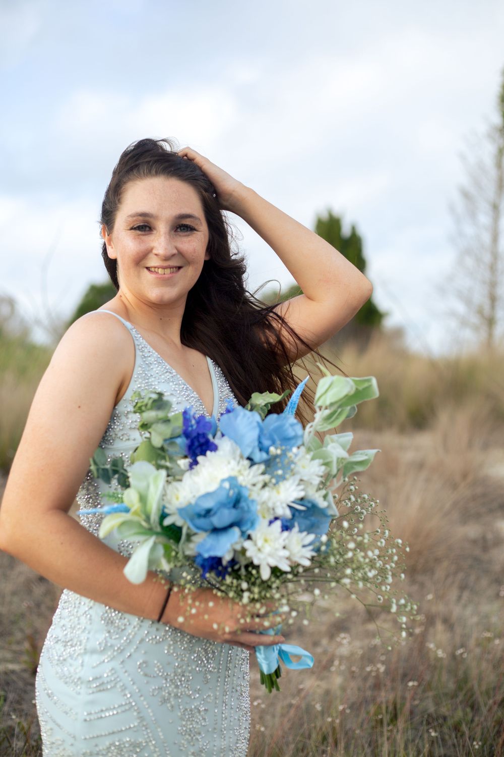 Bride in beaded gown holding blue bouquet while posing in rustic outdoor setting.