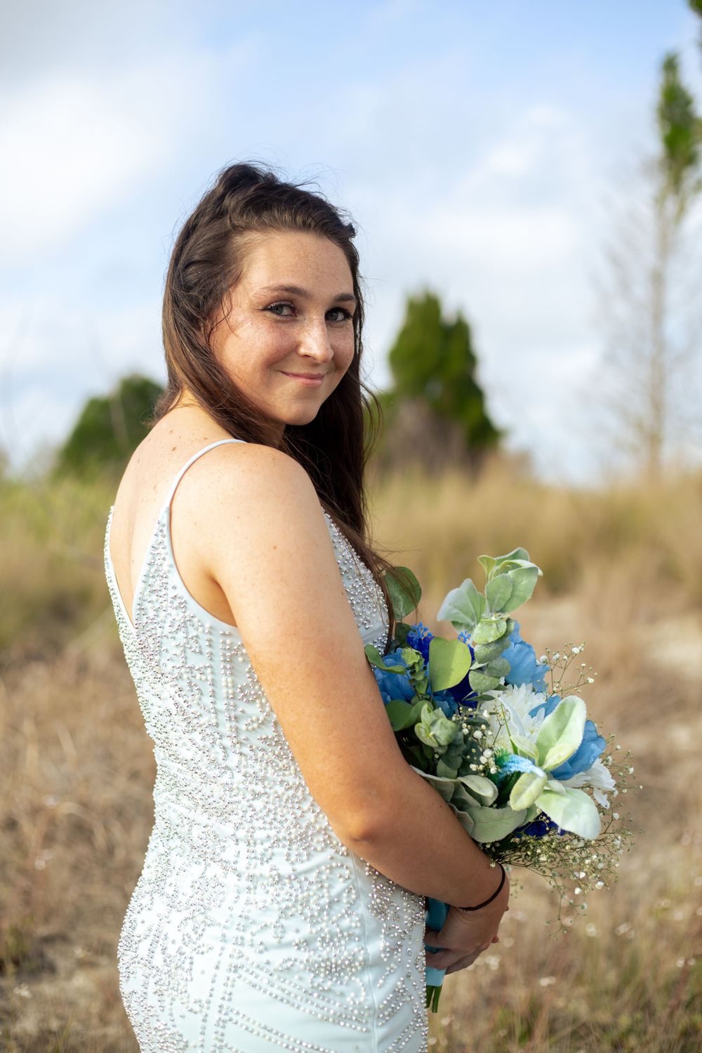 Bride in sparkly dress holding blue and green bouquet in field during golden hour.
