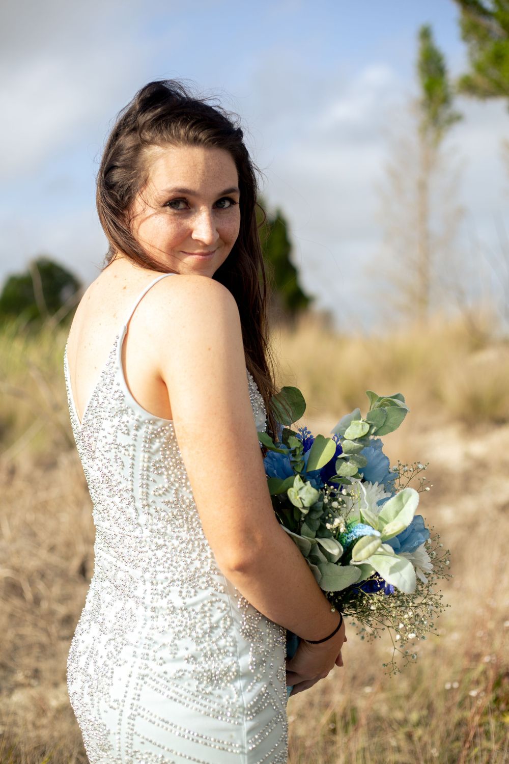 Elegant bridal portrait in sequined dress with blue floral bouquet in natural setting.