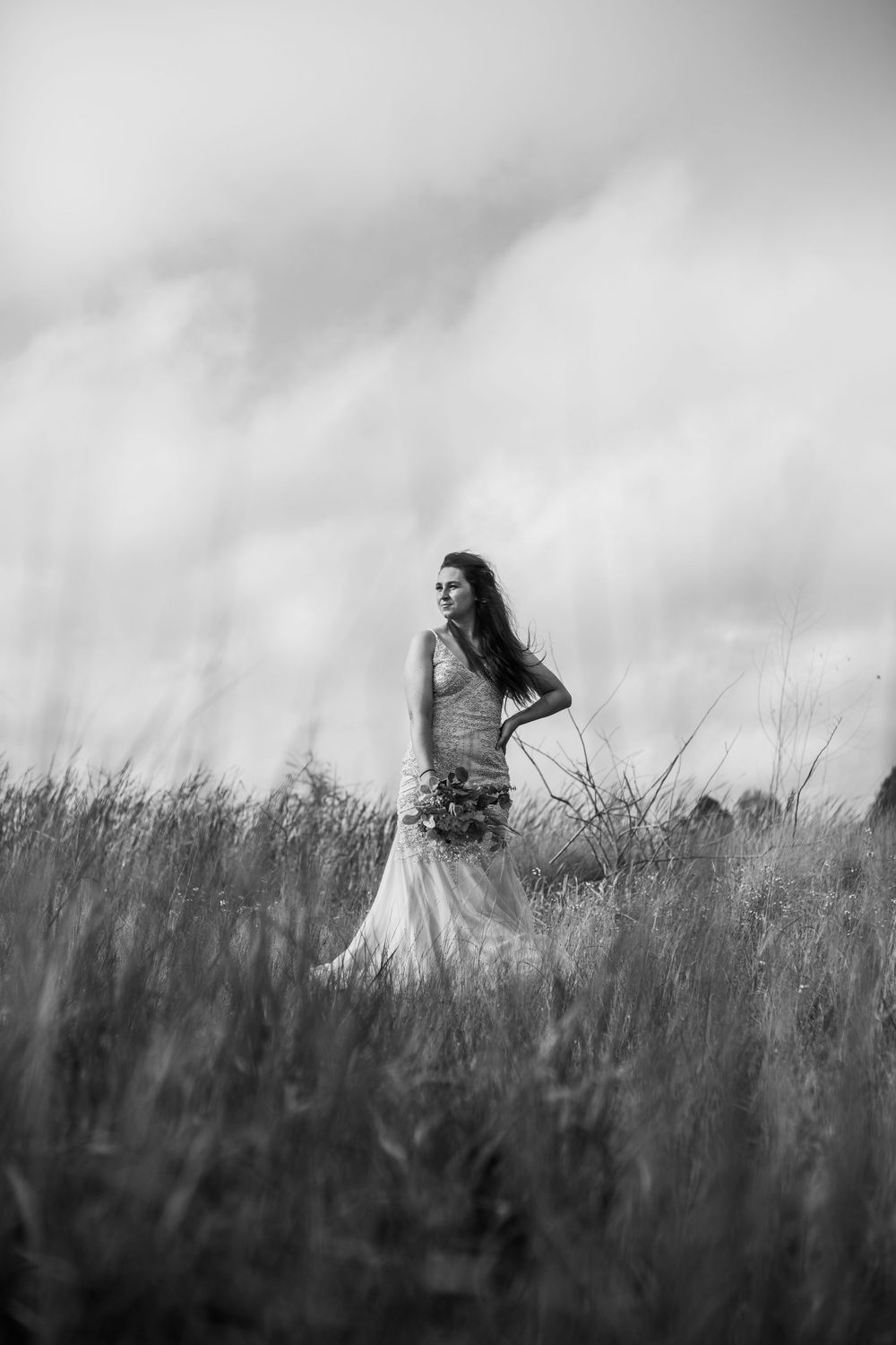 A dramatic black and white portrait of a bride standing in a field of tall grass under cloudy skies.