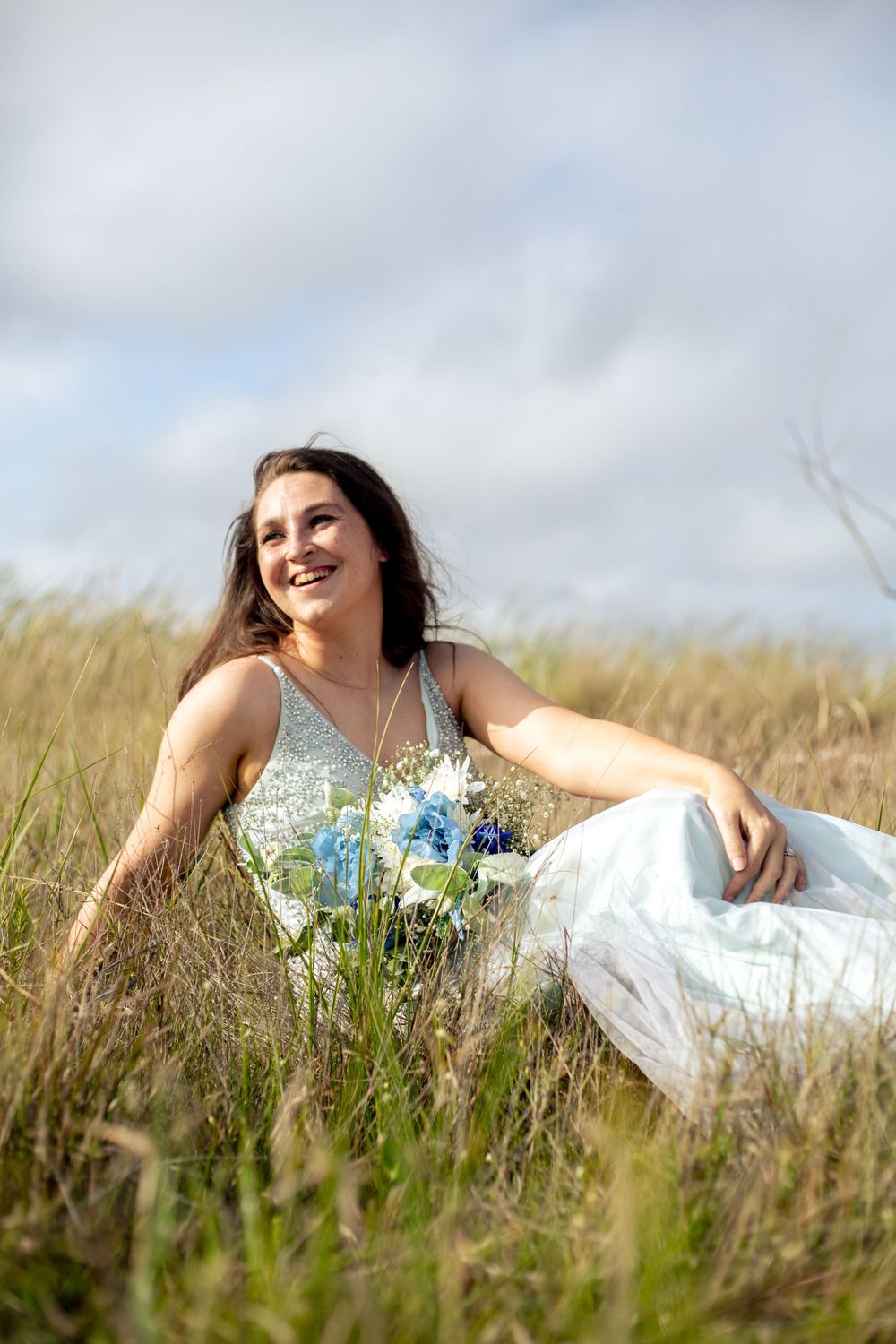 A joyful bridal portrait amidst tall grass with a blue and white bouquet.