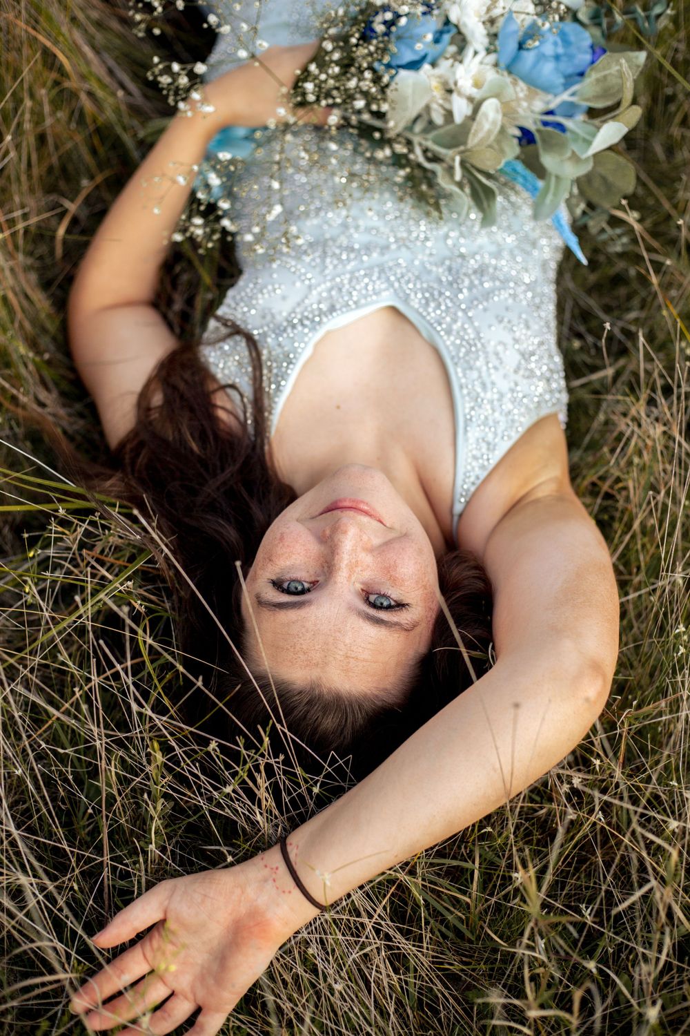 Overhead view of bride lying in grass holding blue and white bouquet.