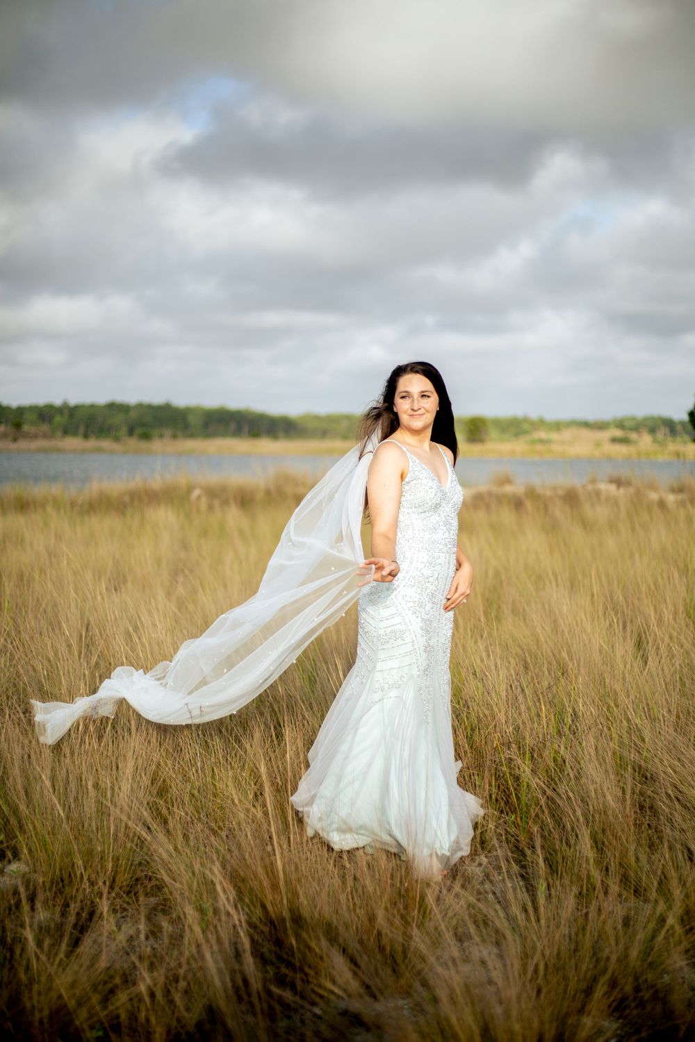Bride in fitted lace gown with flowing veil poses in coastal marsh at sunset.