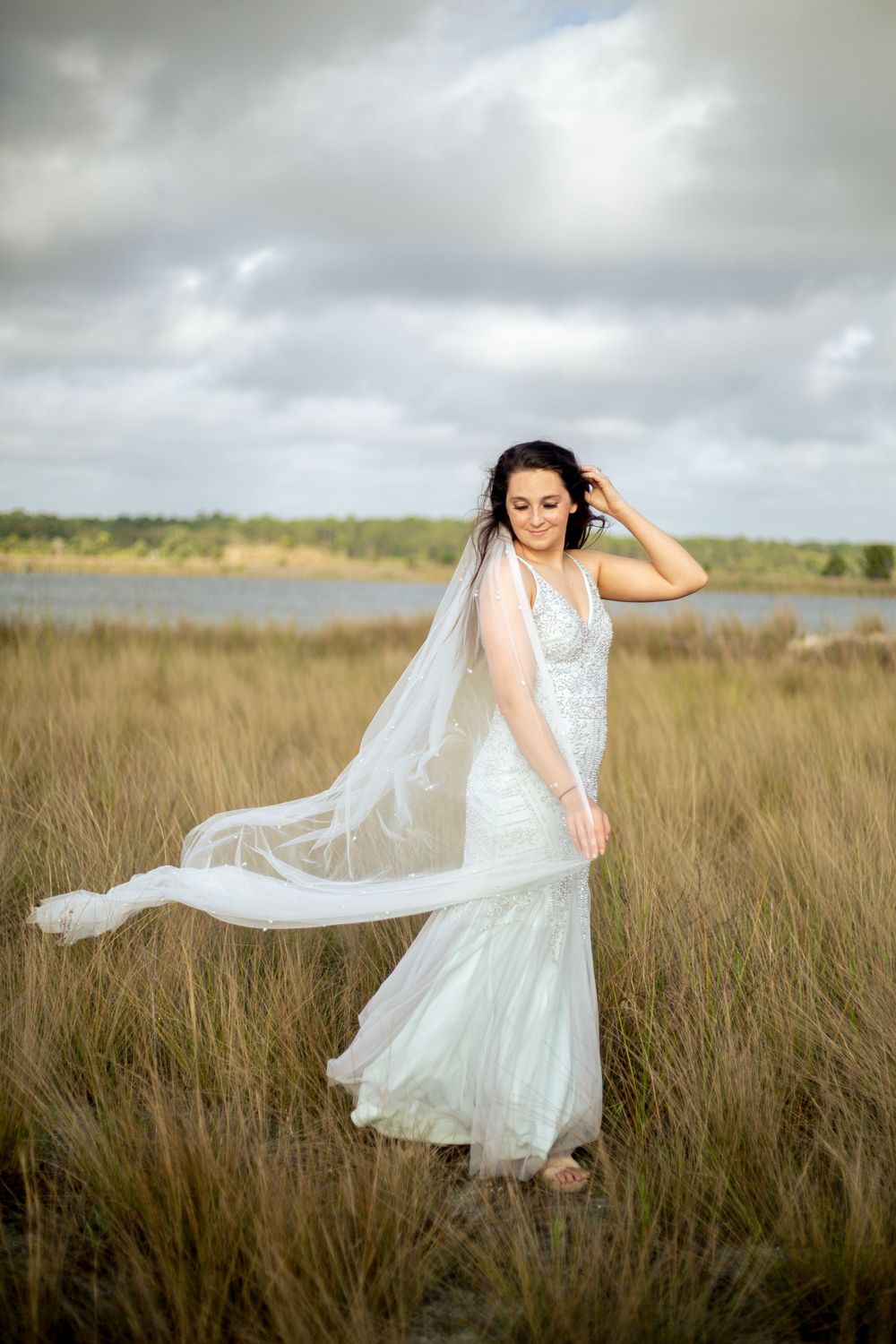 Bride in flowing dress touches her hair while standing in marsh grass near water.