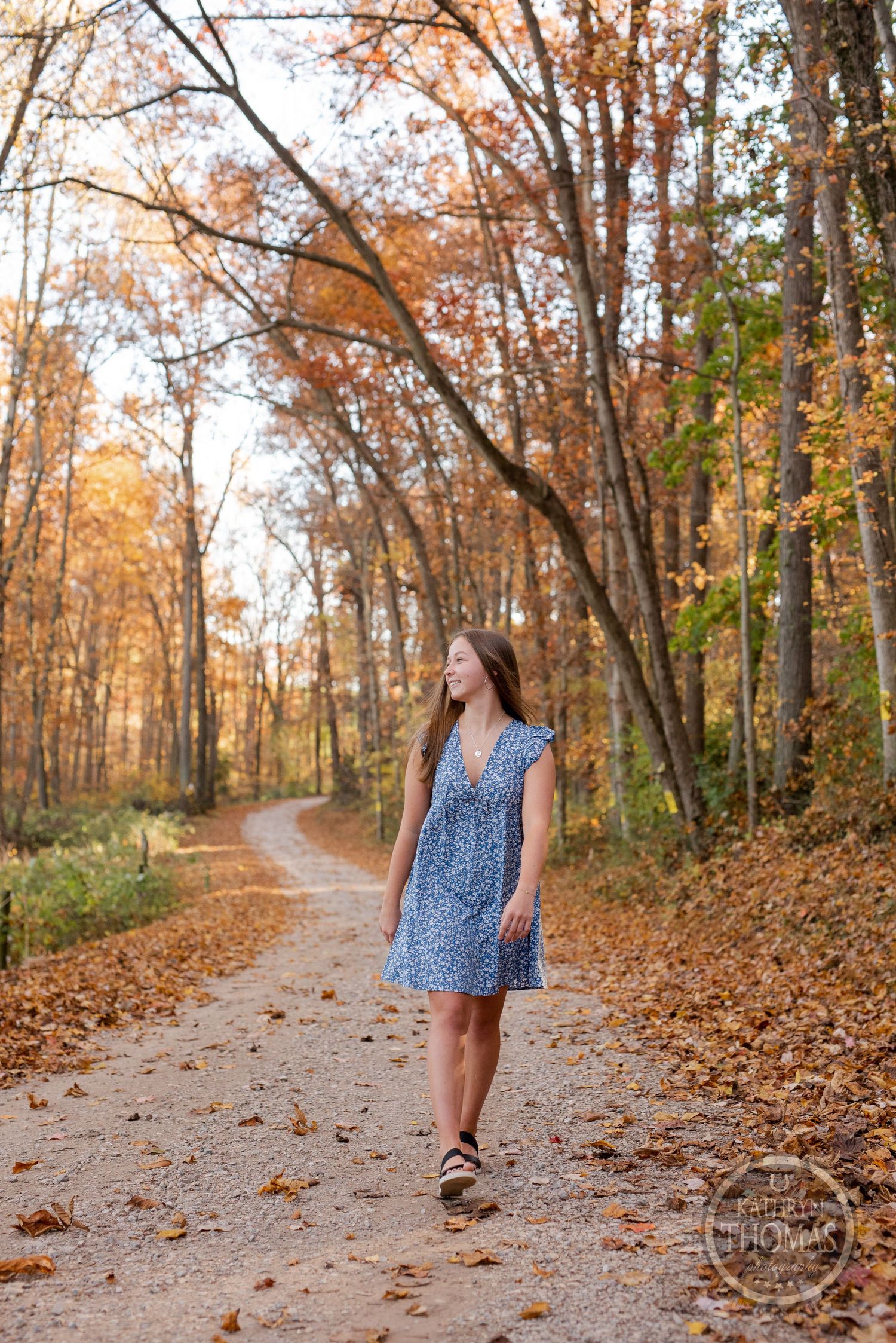 Someone in a blue floral dress walks along a leaf-strewn path through an autumn forest.