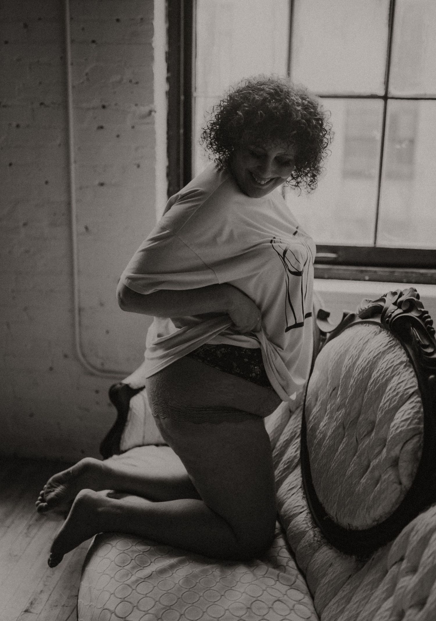 A black and white artistic photograph shows a woman with curly hair posing on a vintage couch near a window.