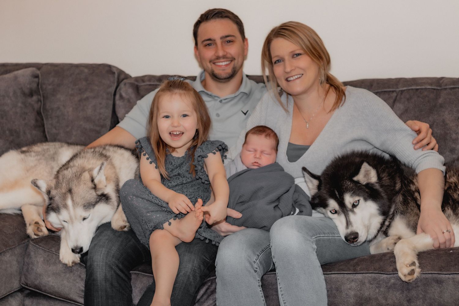Newborn photos. Family relaxing on gray couch with two husky dogs on either side. Southern NH