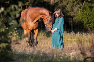 Person in teal dress standing next to brown horse in meadow.