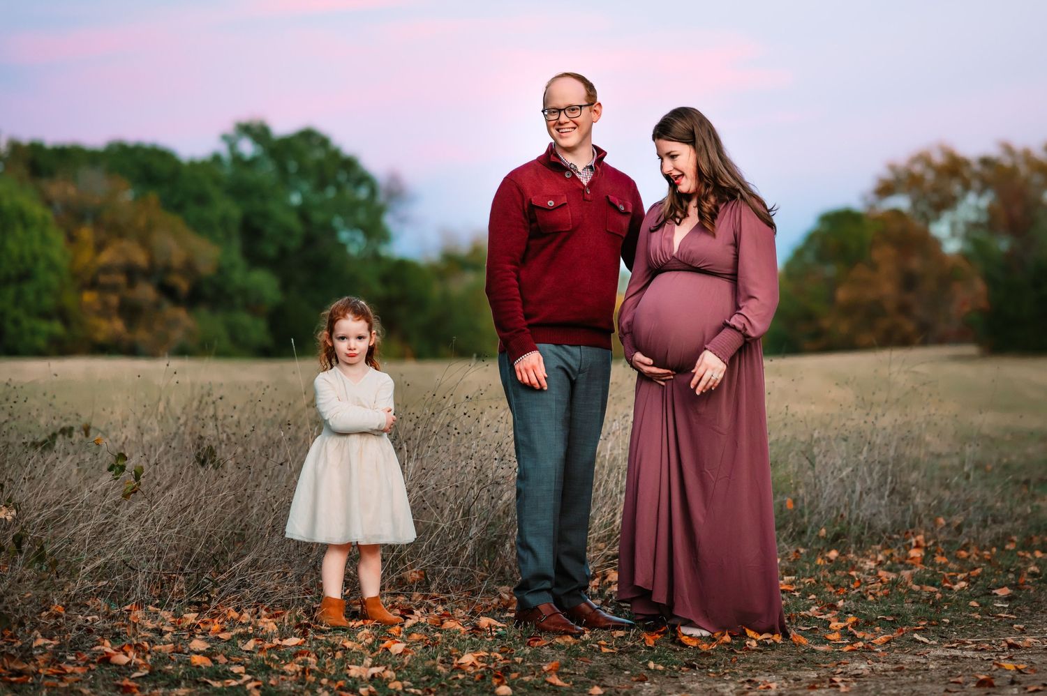 toddler looks at camera crossing arms w/ mom and dad in the background holding mom's baby bump during dallas area maternity session outdoors