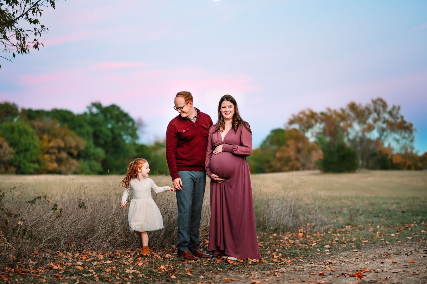 mom in purple dress holds her baby bump while father looks at preschool daughter running around them during dallas maternity photosession