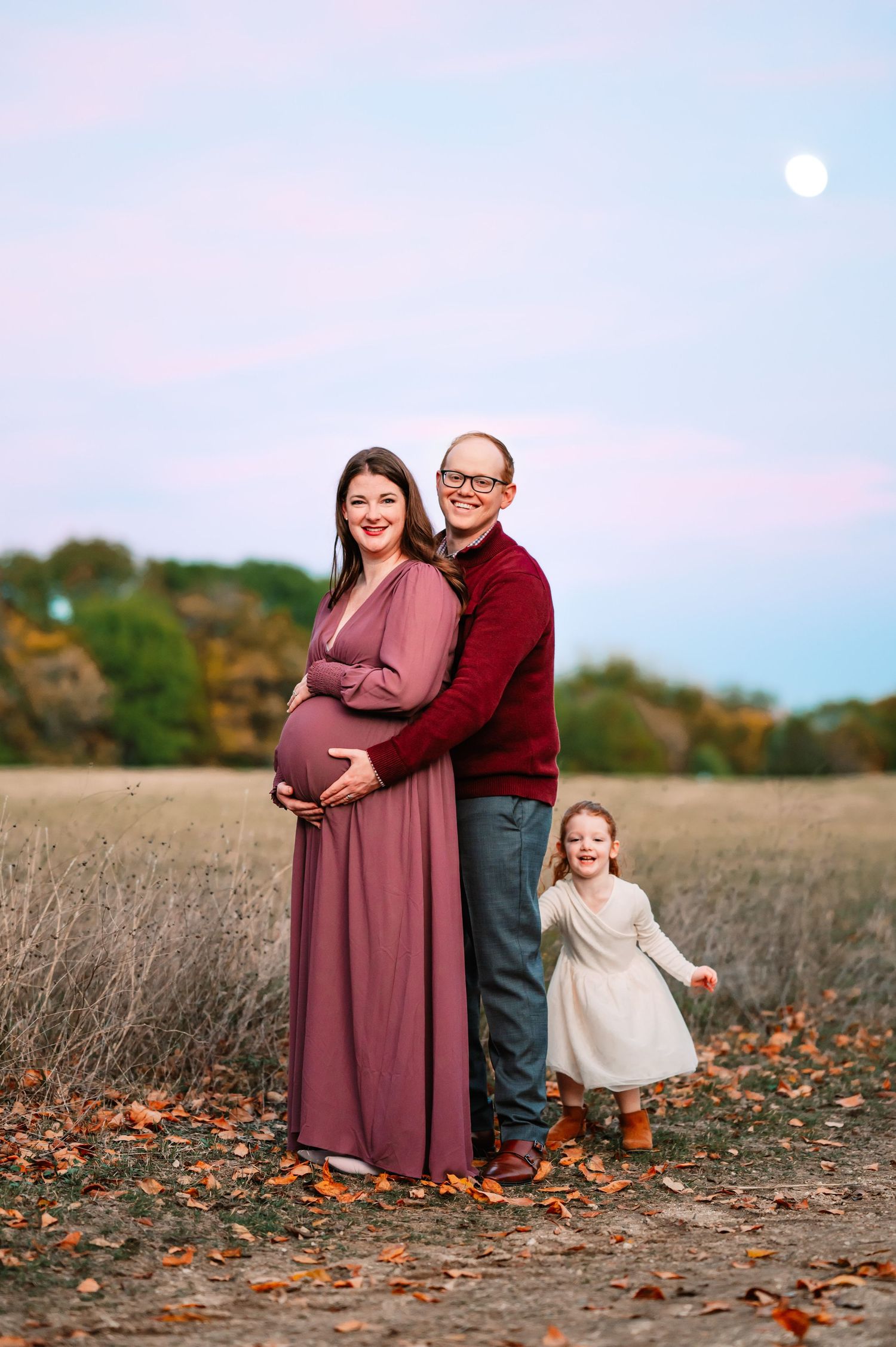 Dad behind mom reaching to hold her baby bump while toddler daughter runs during outdoor Dallas area maternity photo session at sunset