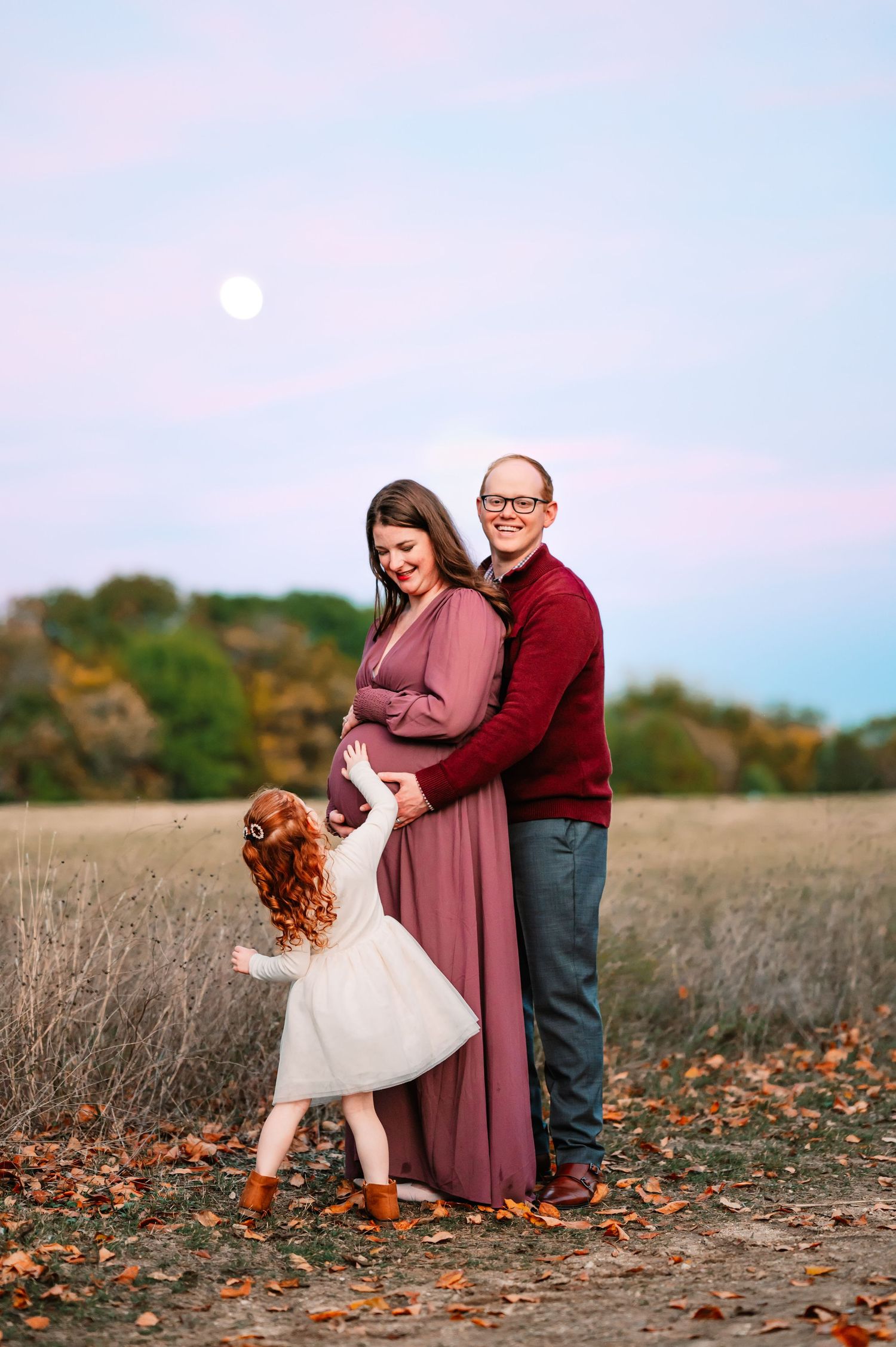 Toddler reaches for mom's baby bump during outdoor fall maternity photo session at sunset