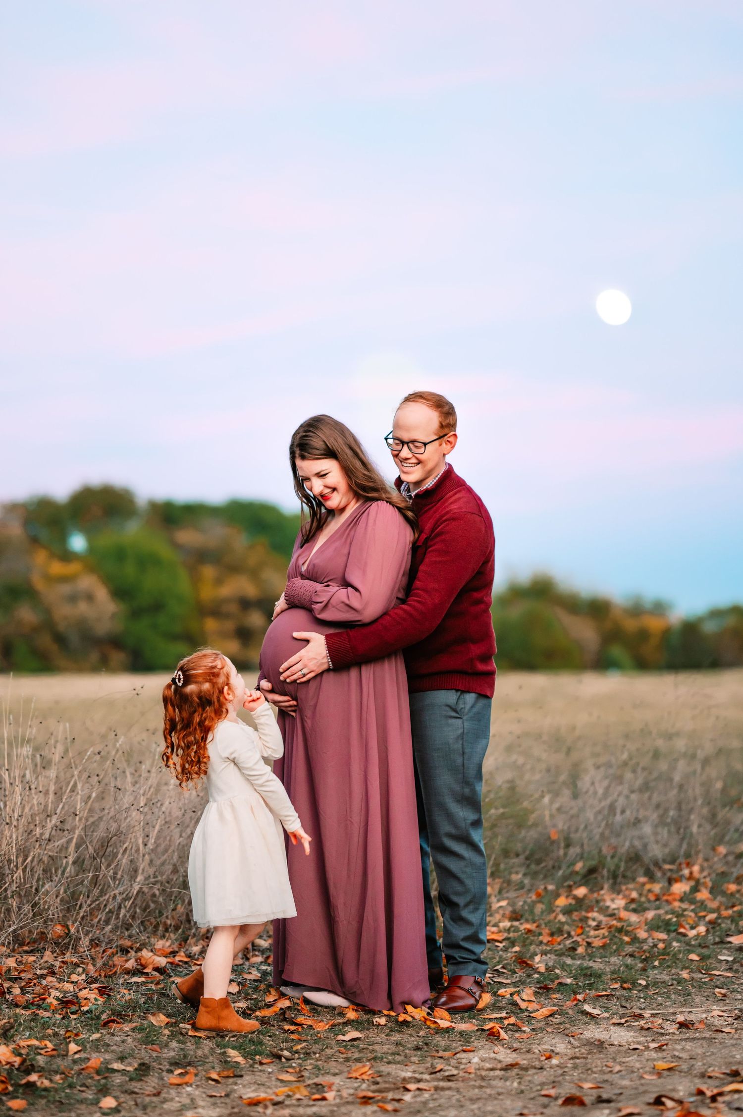 Toddler reaches for mom's baby bump during outdoor fall maternity photo session at sunset